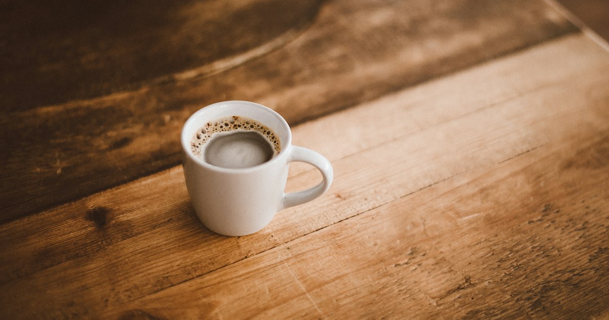 A white cup of coffee left on a warm, worn wooden table