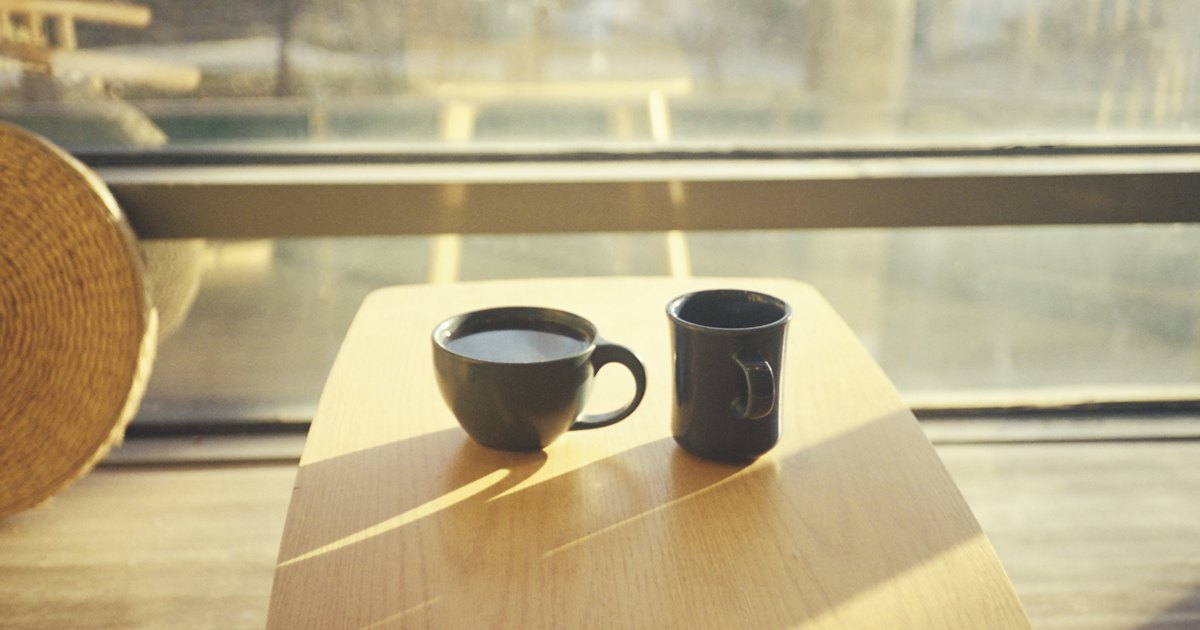 2 cups of coffee side by side on a wooden table by a sunlit window