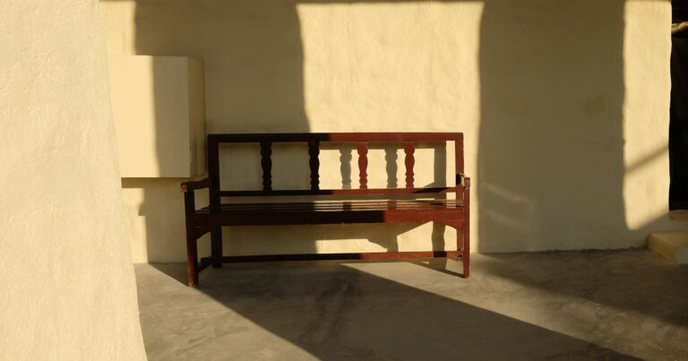 An empty wooden bench against a cream wall, half in warm sunlight and half in shadow