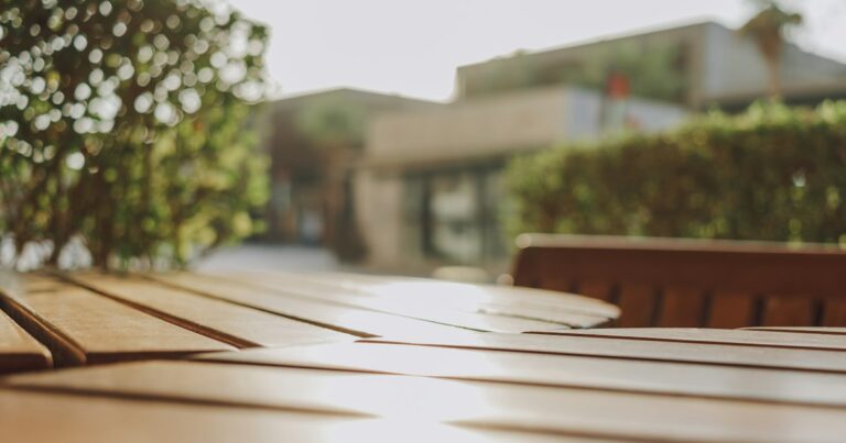 An empty wooden café table in warm sunlight, with green hedges and a low building softly blurred behind it