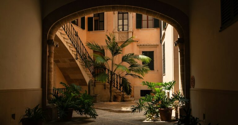 A stone archway opening onto a quiet sunlit courtyard with an outdoor staircase, a palm and potted plants