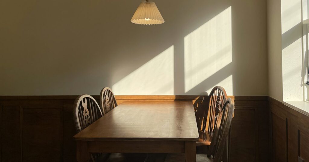 An empty wooden table and chairs under a paper lampshade, with afternoon sunlight falling across the wall