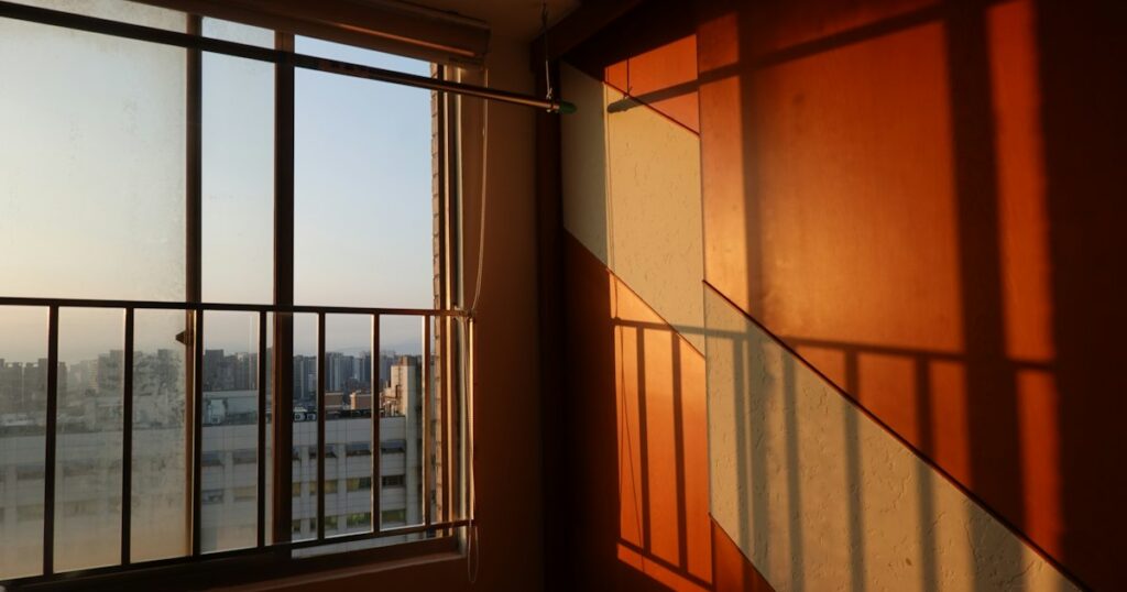 Evening sun through an apartment window casting warm railing shadows on the wall, with a city skyline outside