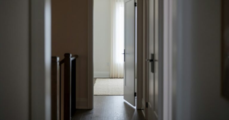 A quiet hallway leading to a half-open door and a bright room with a sheer curtain beyond