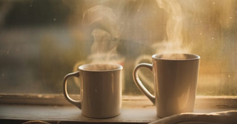 2 steaming mugs on a windowsill in warm early light, with a soft blanket beside them