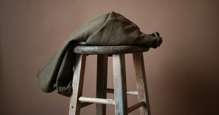 An olive jacket draped over an old wooden stool against a warm terracotta wall