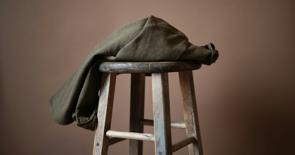 An olive jacket draped over an old wooden stool against a warm terracotta wall