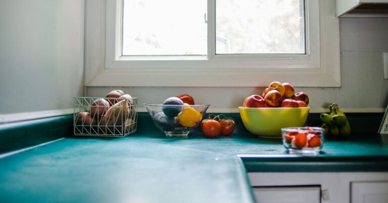 A bright kitchen counter under a window with bowls of apples, tomatoes and other fruit