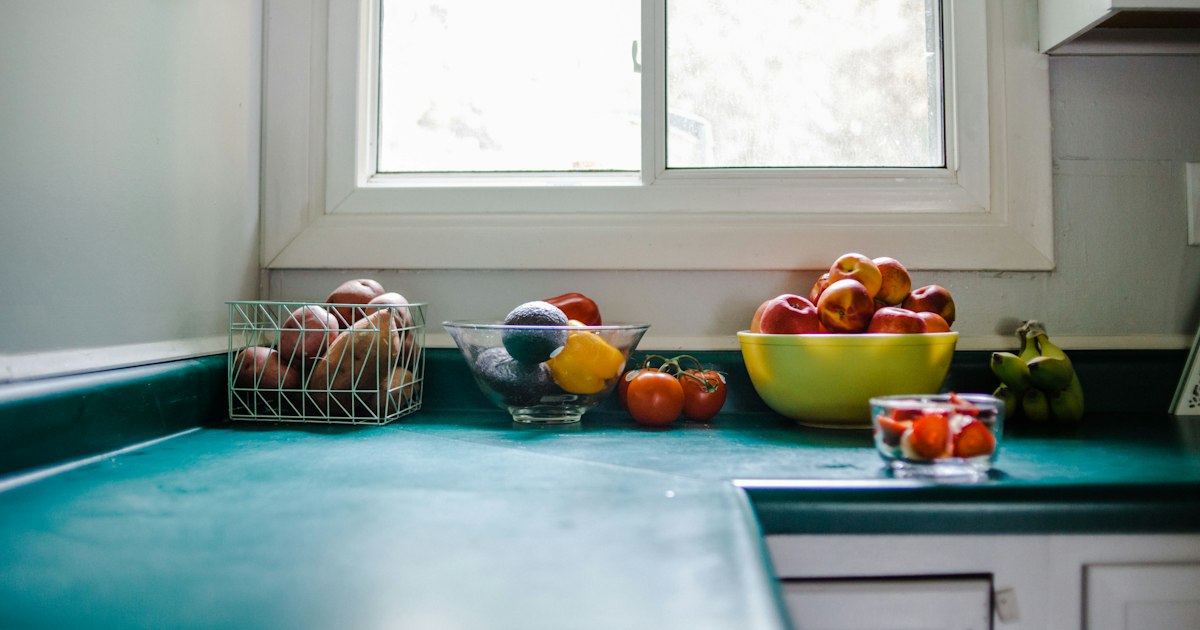 A bright kitchen counter under a window with bowls of apples, tomatoes and other fruit