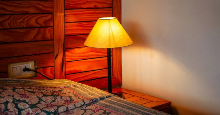 A table lamp with a yellow shade glowing beside a bed with a patterned cover against a wooden headboard