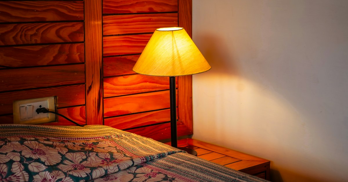 A table lamp with a yellow shade glowing beside a bed with a patterned cover against a wooden headboard