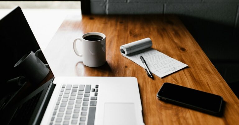 A laptop, a mug of coffee, a notepad with a pen and a phone on a warm wooden desk