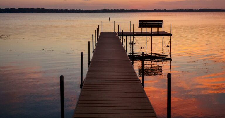 A wooden dock leading out to an empty bench over a calm lake at sunset
