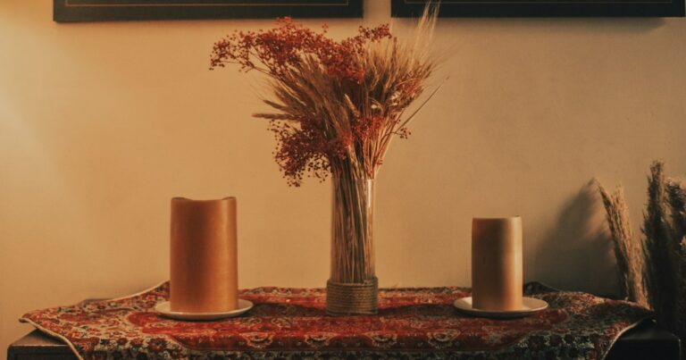 Dried flowers in a glass vase between 2 candles on a patterned cloth in warm light