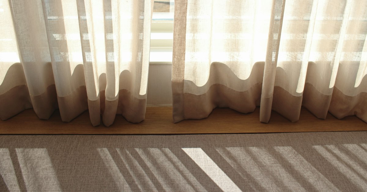 Sheer cream curtains glowing with daylight, casting soft striped shadows across a pale floor