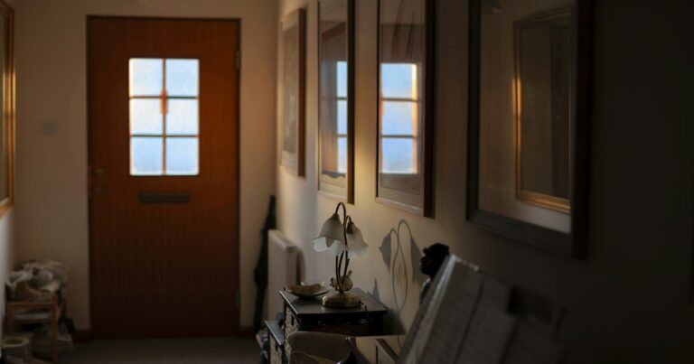 A front door seen from inside a warm hallway with a small lamp and framed pictures