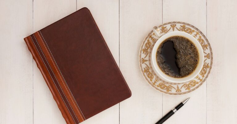 A closed leather notebook, a cup of black coffee and a pen on a pale wooden table