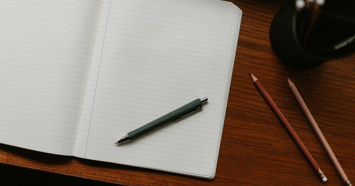 An open lined notebook with a green pen resting on it, beside 2 pencils on a warm wooden desk
