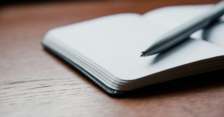An open blank notebook with a silver pen resting on it, on a warm wooden table