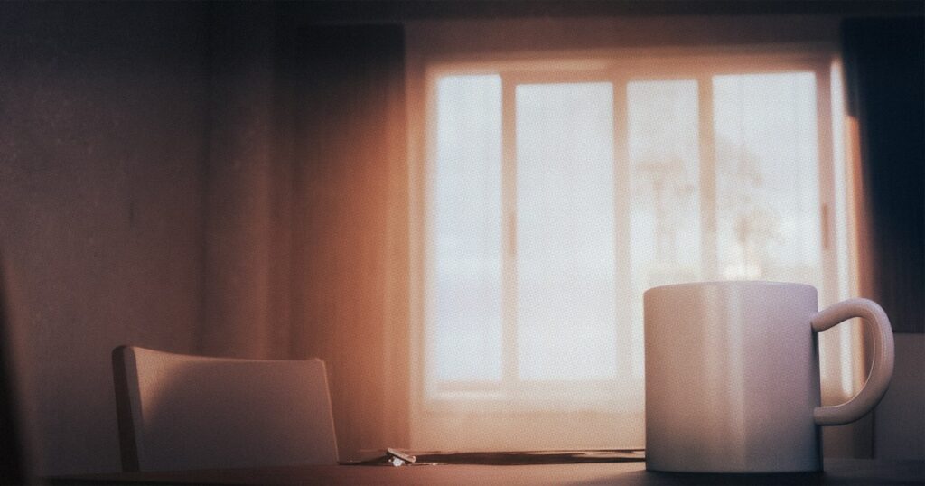 A white mug on a table beside an empty chair, in front of a window glowing with soft morning light