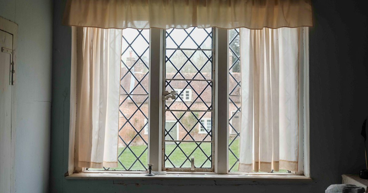 A leaded window with sheer cream curtains looking out onto a lawn and an unfamiliar brick house