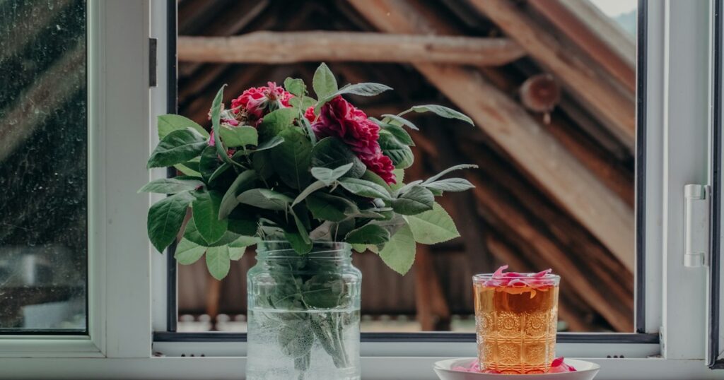 Garden roses in a glass jar and a glass of tea on a windowsill, with a wooden barn outside