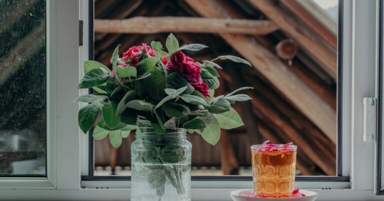 Garden roses in a glass jar and a glass of tea on a windowsill, with a wooden barn outside