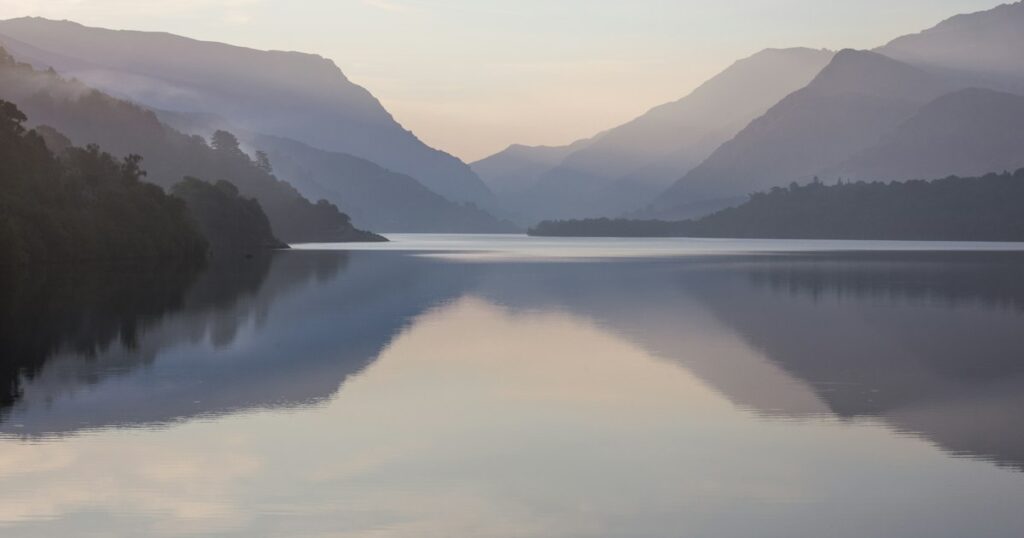 A still lake reflecting misty hills in soft early morning light