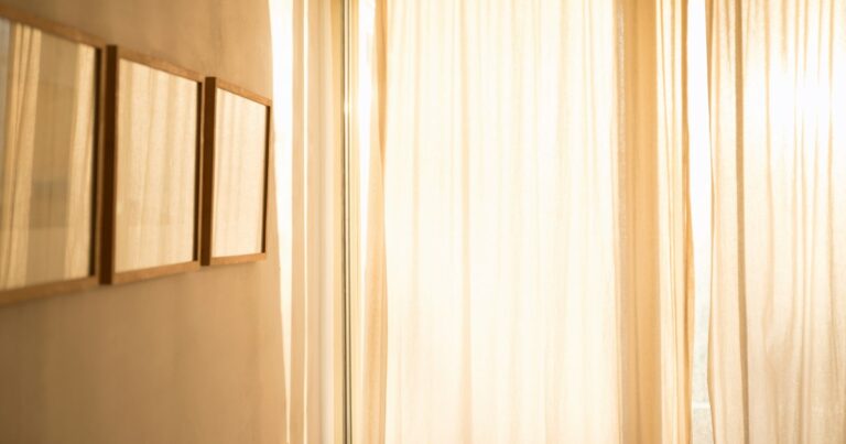 Bright sunlight through pale linen curtains beside 3 empty wooden picture frames on a cream wall