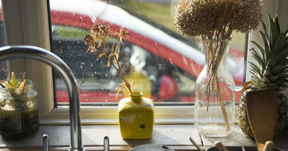 A sunlit kitchen windowsill with a small yellow vase, dried flowers in a glass carafe and a pineapple