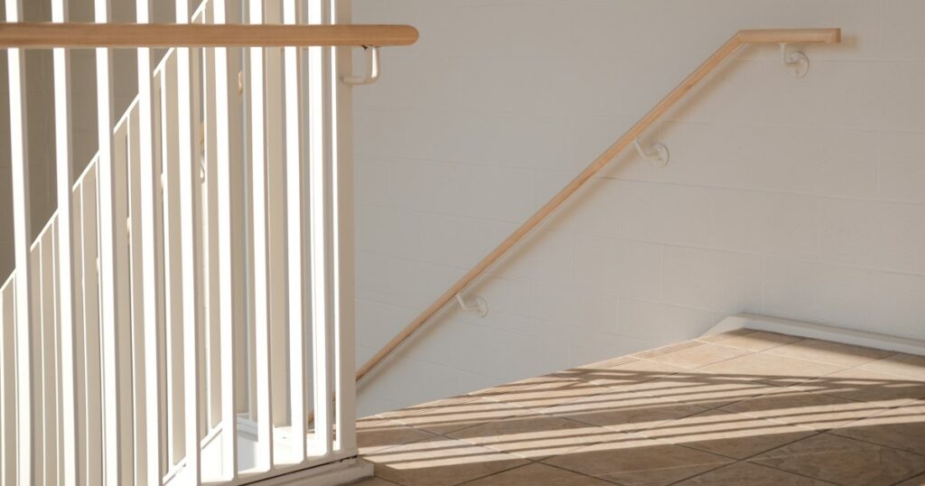 Sunlight falling across a pale stairwell with a wooden handrail leading down to a lower landing