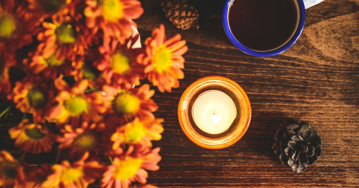 A lit tealight among orange flowers and pine cones on a wooden table, seen from above
