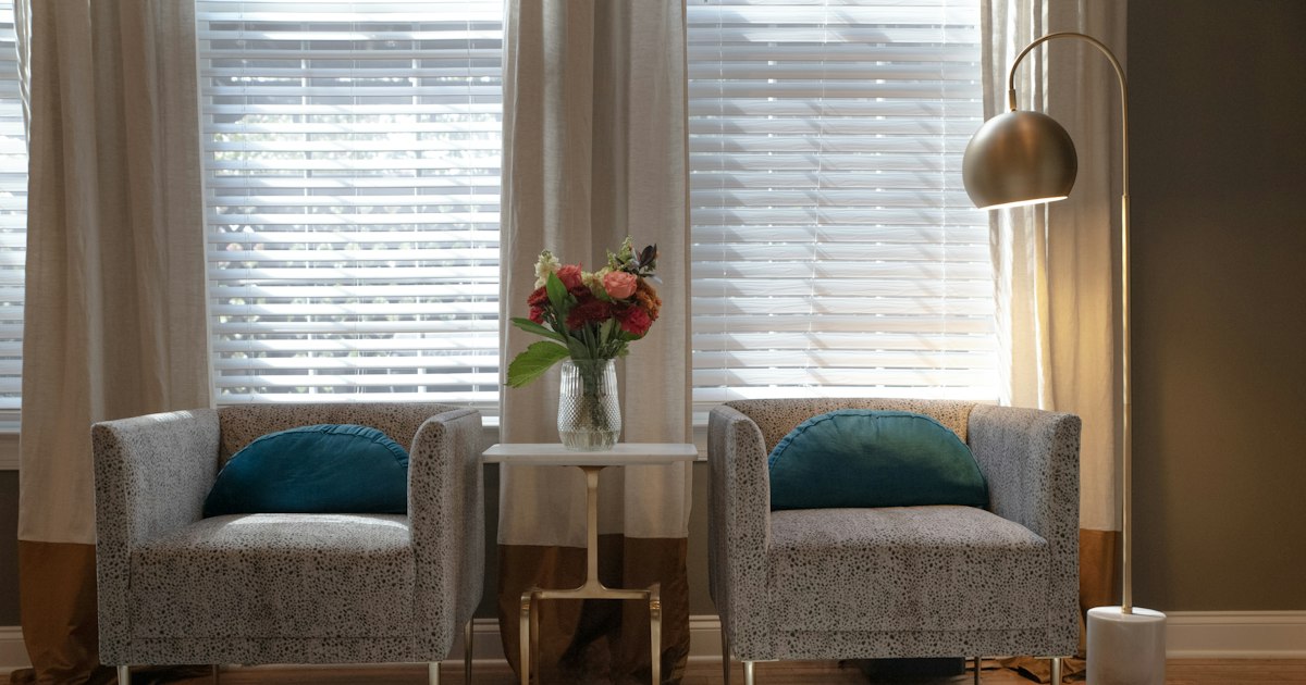 Two grey armchairs with teal cushions either side of a small table with flowers, in front of a window with blinds
