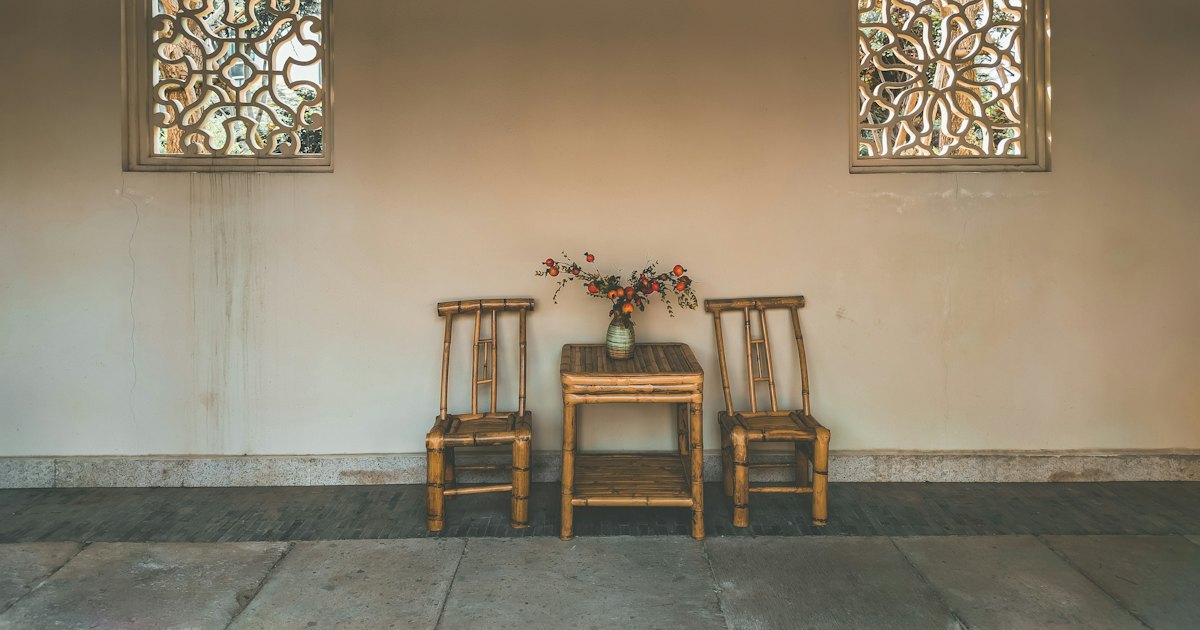Two bamboo chairs and a small table with a posy against a pale wall