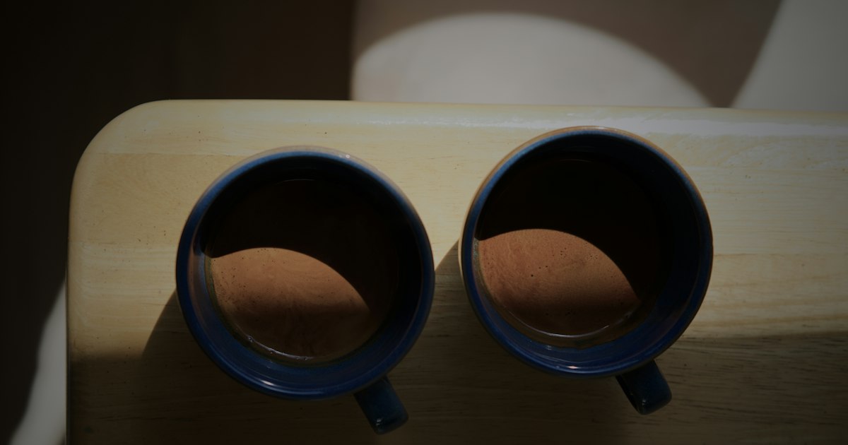 Two blue cups of coffee side by side on a wooden table, sharing a patch of warm sunlight