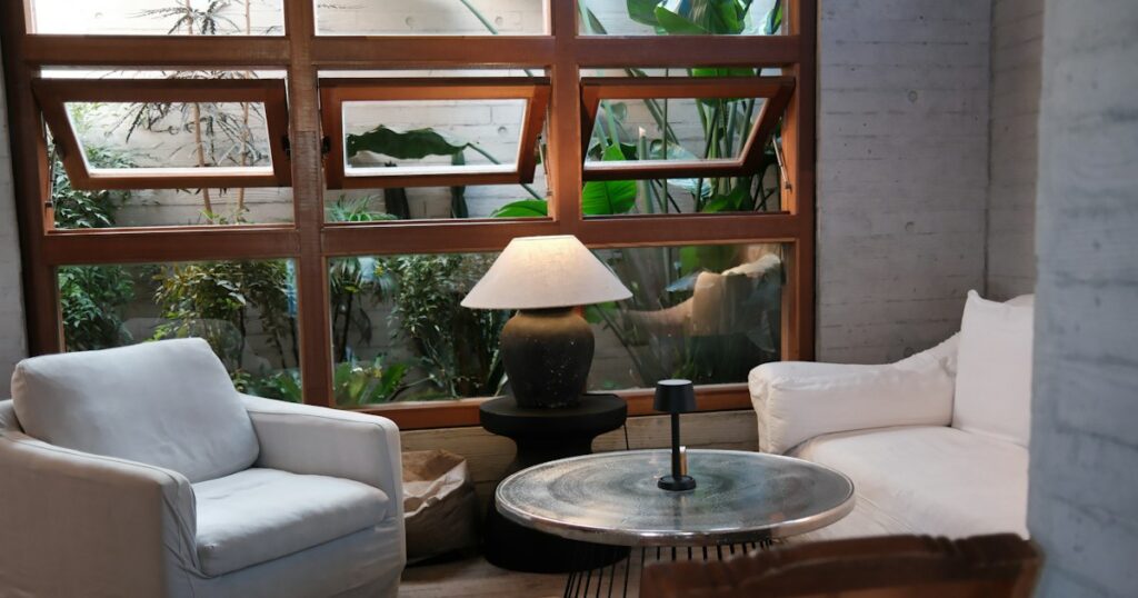 A white armchair and a white sofa facing each other across a round low table, in front of a wood-framed window full of green plants