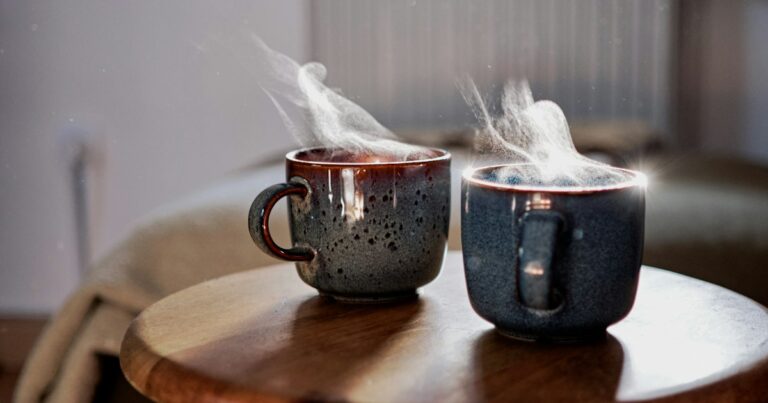 Two steaming speckled ceramic mugs side by side on a small round wooden table in soft daylight