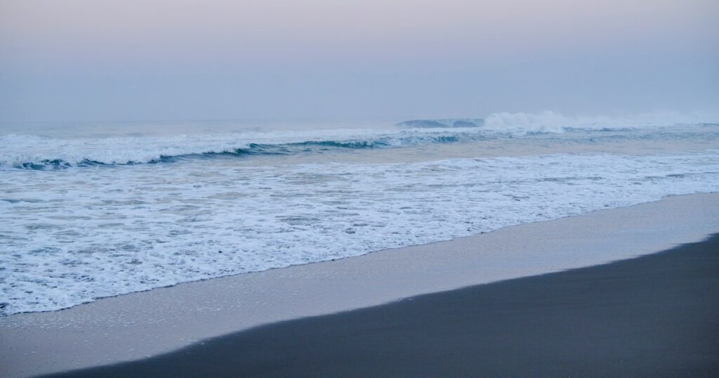 A wave breaking and running up a quiet sandy beach under a soft pink dawn sky