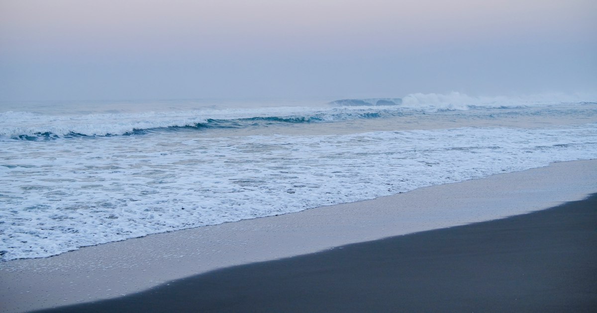 A wave breaking and running up a quiet sandy beach under a soft pink dawn sky
