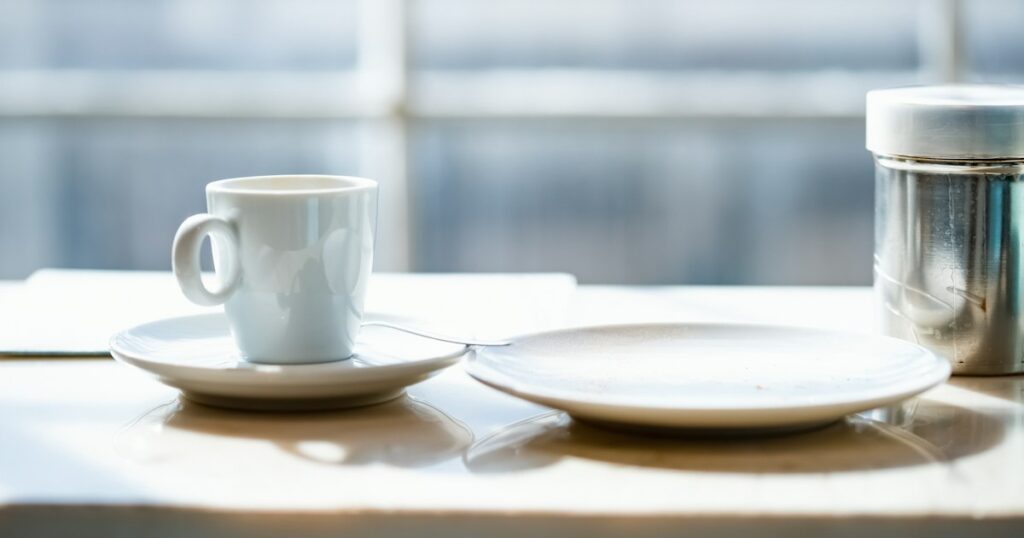 A white cup on a saucer beside an empty plate and a metal tin on a sunlit table by a bright window