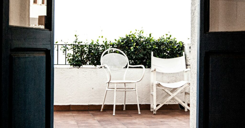Two mismatched white chairs a little apart on a bright balcony, seen through an open door