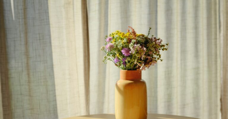 A bunch of wildflowers in a yellow vase in front of sunlit linen curtains